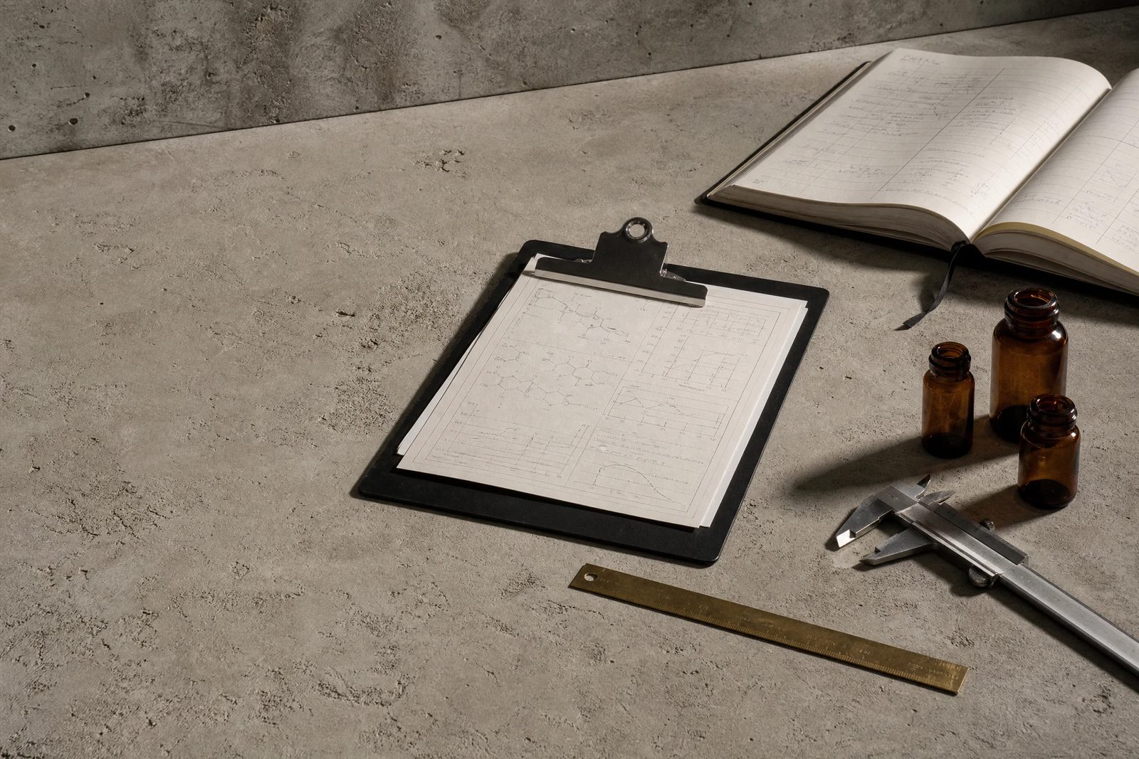 Clipboard of structural diagrams, a bound lab notebook, amber vials and brass calipers on a concrete surface